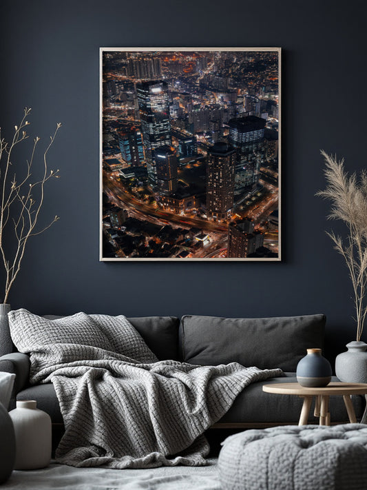 A framed vertical art print of a city skyline at night with glowing lights, displayed on a dark navy wall above a gray sofa in a styled living room.