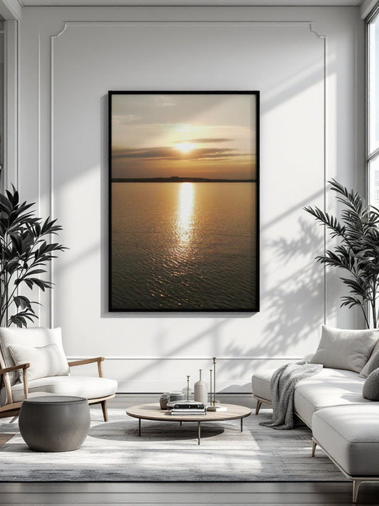 A framed vertical canvas print of a golden sunset over water, displayed on a white paneled wall in a modern, sunlit living room with a sofa, armchair, and coffee table.