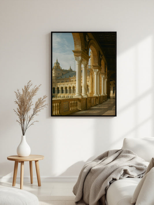 A framed art print depicting a classical architectural colonnade with arches and a building in the background, displayed on a white wall in a modern living room setting next to a small wooden side table with a vase of dried grass and a white sofa with a gray throw blanket.