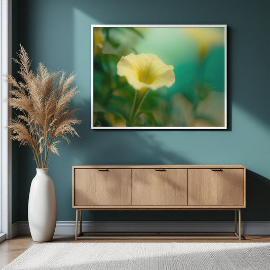 A framed wall art print showing a yellow flower on a soft green background, displayed above a light wood console table in a styled room with a white vase of dried grass against a dark teal wall.
