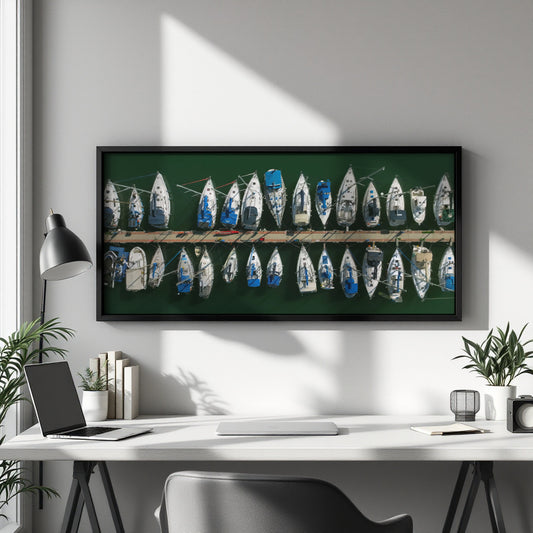 A framed horizontal canvas print of vintage sailboats on a green background, displayed on a light gray wall above a white desk in a modern home office setting.