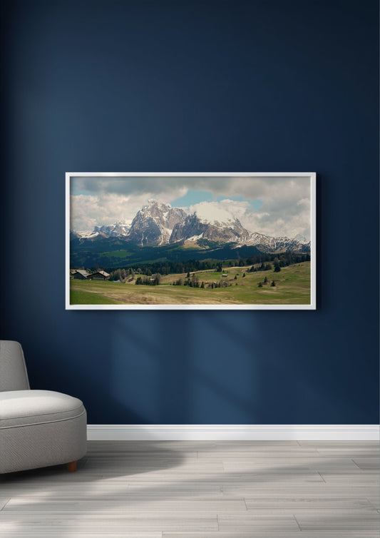 A framed landscape art print of snow-capped mountains and green valleys, displayed on a dark navy blue wall above a light gray floor, with a partial view of a gray armchair on the left.