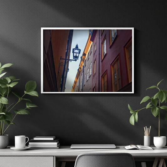 A framed vertical art print depicting a narrow European street with red and orange buildings, hanging on a dark gray wall above a styled desk with plants and books.