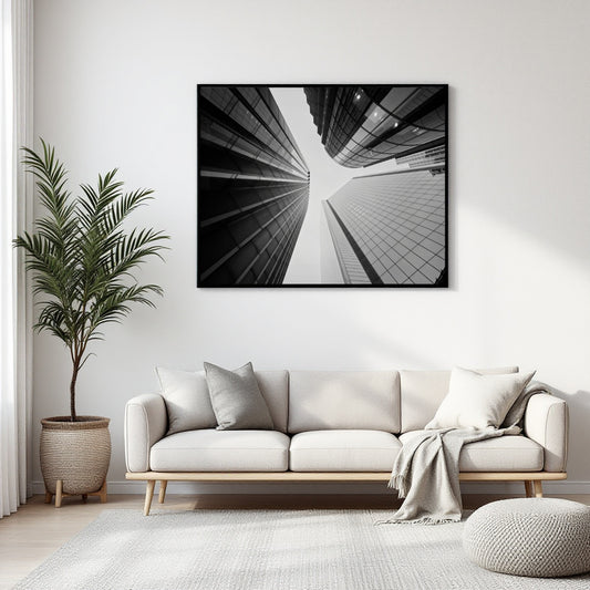 A black and white architectural canvas print of modern skyscrapers hangs on a white wall above a light beige sofa in a modern living room setting with a potted palm and neutral decor.