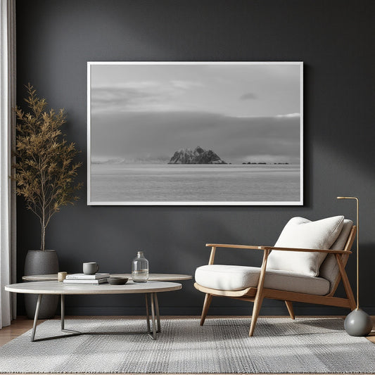 A large black and white framed landscape art print of a rocky island on the sea, displayed on a dark gray wall in a modern living room setting with a wooden armchair, round coffee table, and a potted plant.