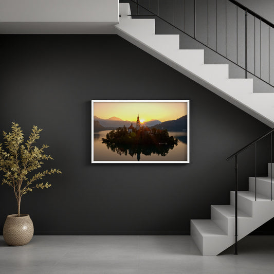 A framed landscape art print of Bled Island in Slovenia, showing a church on an island reflected in the water at sunset, displayed on a dark gray wall in a modern interior setting with a staircase and a potted plant.