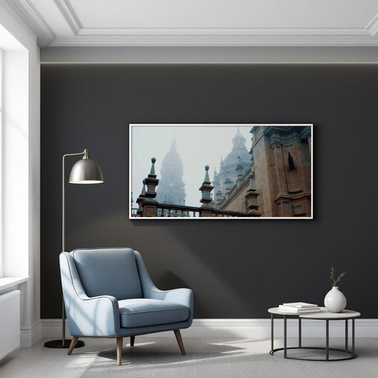 A framed panoramic art print of historic European architecture with domes and towers, displayed on a dark charcoal wall in a modern living room setting with a blue armchair, floor lamp, and side table.