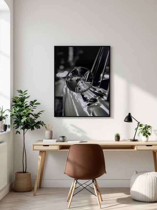 A black and white framed wall art print depicting a close-up of a car side mirror and chrome details, displayed in a modern, sunlit home office setting above a wooden desk and brown chair.