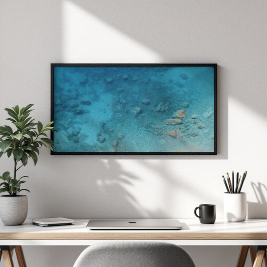 A framed wall art print showing an underwater ocean scene in shades of blue and turquoise, hung on a white wall above a desk with a plant, laptop, mug, and pencils.