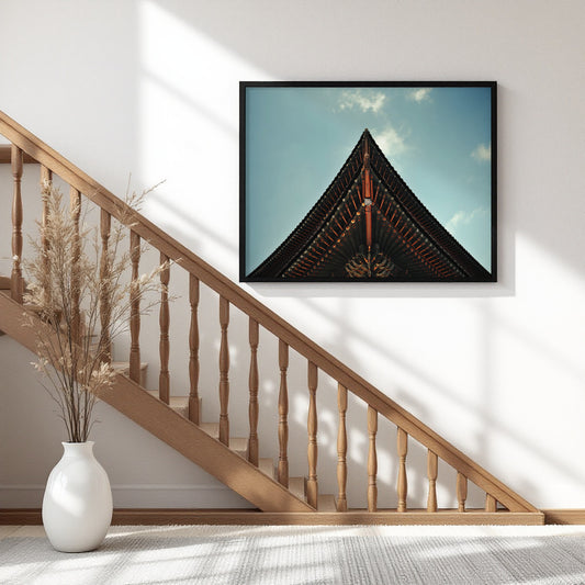 A framed wall art print showing the pointed dark roof of a traditional building under a blue sky, displayed above a wooden staircase in a bright modern interior with a white vase of dried grass on the floor.
