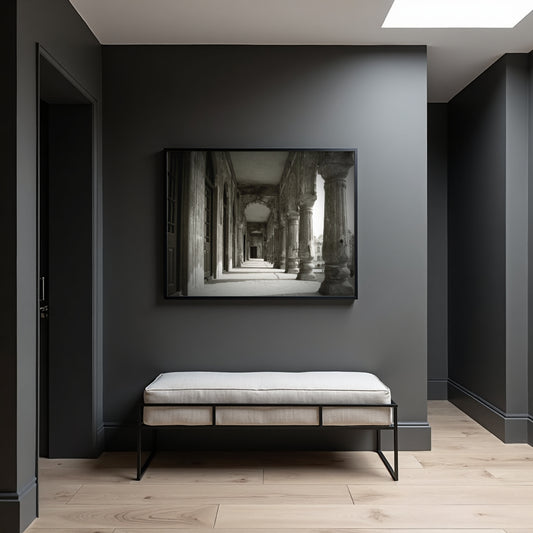 A black-framed black and white art print of an ancient stone corridor hangs on a dark gray wall above a light beige upholstered bench with black metal legs in a modern, minimalist room.
