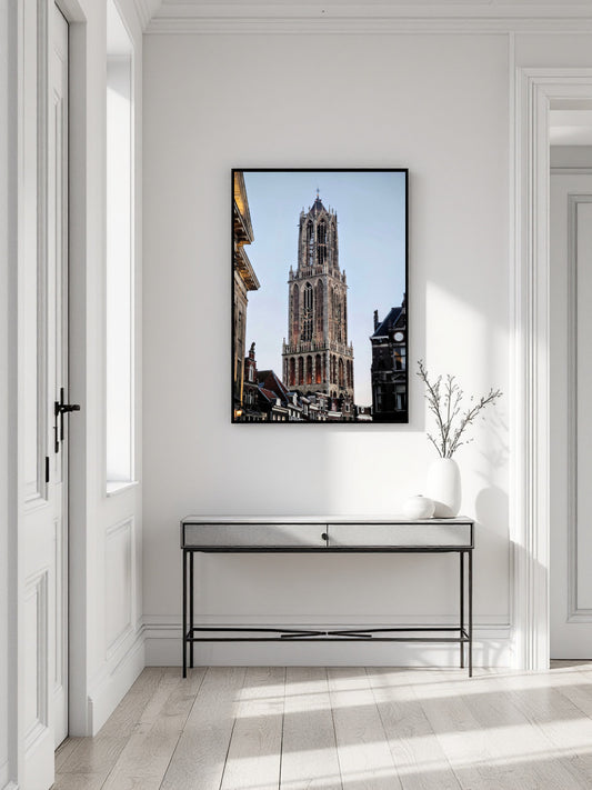 A framed vertical art print of the Belfry of Brussels under a clear blue sky, displayed on a white wall above a modern console table in a bright, minimalist hallway.
