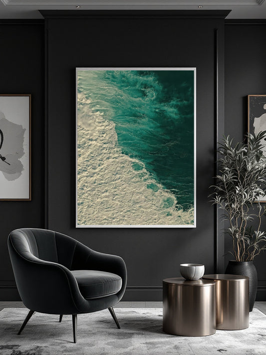 A vertical framed art print featuring an aerial view of turquoise ocean waves and white sea foam, displayed on a dark charcoal wall in a modern living room setting with a black armchair, metallic side tables, and a potted plant.