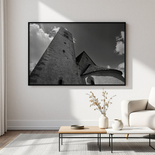 A large black and white framed wall art print depicting a stone tower and building facade under a cloudy sky, displayed in a modern living room setting above a low wooden coffee table and next to a white armchair.