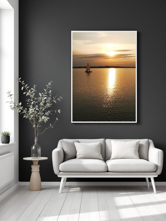 A framed vertical wall art print showing a sunset over calm water with a small sailboat, displayed above a light gray sofa in a modern living room setting.