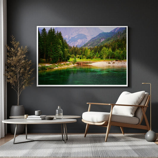 A framed landscape art print depicting a scenic mountain lake with a pine forest and sandy shore, displayed on a dark gray wall in a modern living room setting with a wooden armchair, round coffee table, and potted plant.