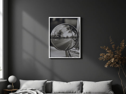A framed black and white art print depicting a car side-view mirror reflecting a road and trees, displayed on a dark gray wall above a modern sofa in a styled living room.