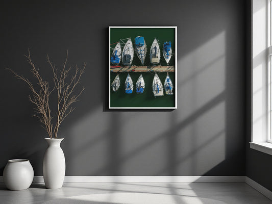 A framed wall art print showing a row of blue and white boats on a dark green background, displayed on a dark gray wall in a modern room setting with a window, vase, and dried branches.