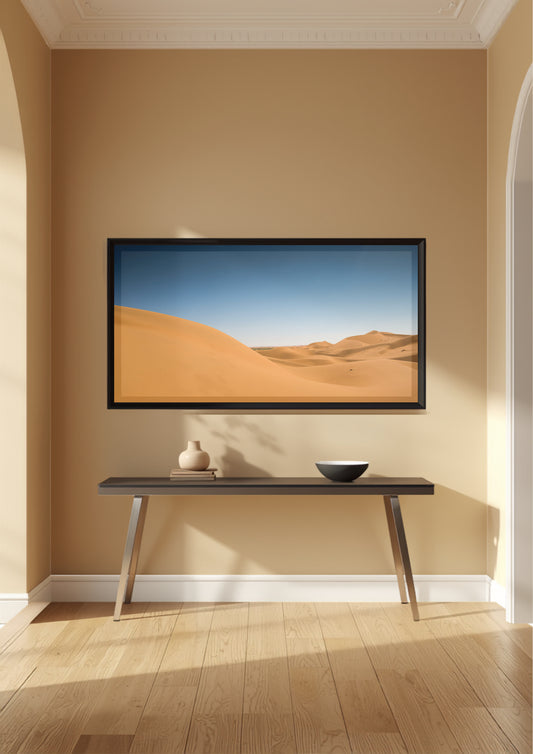 A framed landscape art print depicting golden sand dunes under a blue sky, displayed on a beige wall above a dark console table in a modern, sunlit room with light wood floors.
