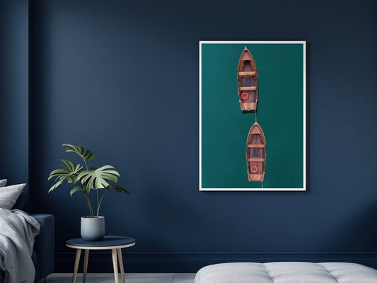 A framed vertical art print featuring two small wooden rowboats on a deep teal-green background, displayed on a dark navy blue wall in a modern bedroom setting next to a potted plant and bed.