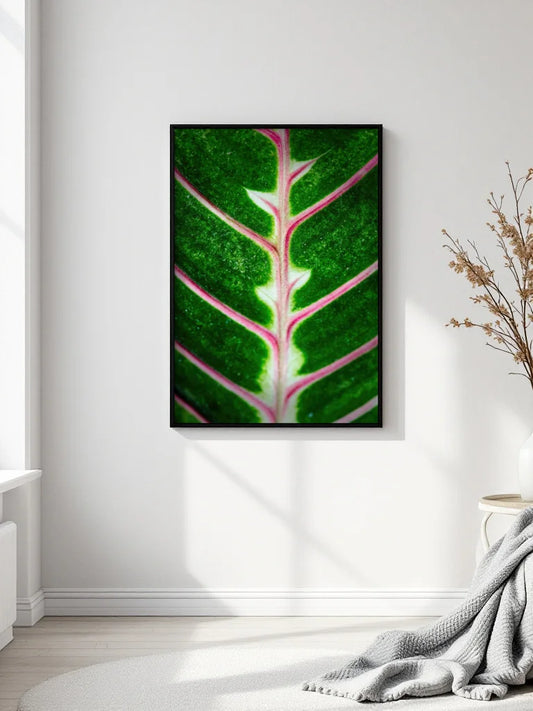 A vertical framed wall art print featuring a close-up green leaf with pink veins, displayed on a white wall in a bright room with a gray throw blanket and dried branches nearby.