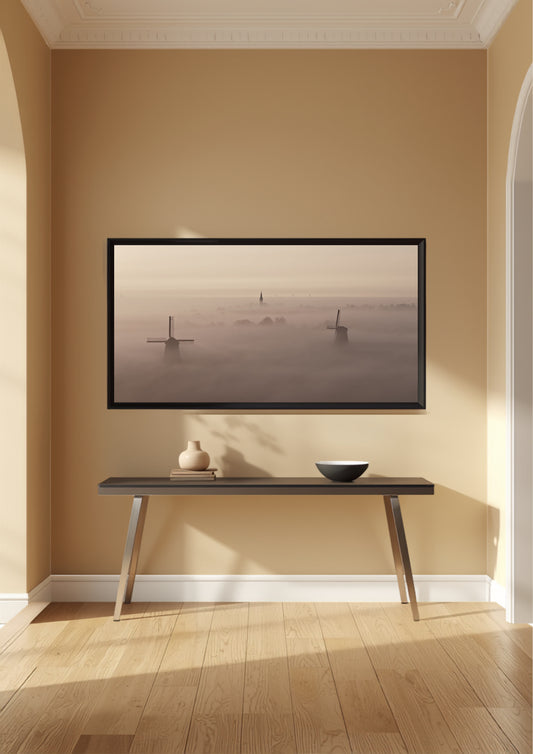 A framed landscape art print depicting windmills in a misty field, displayed on a beige wall above a dark console table in a modern, sunlit room with light wood floors.