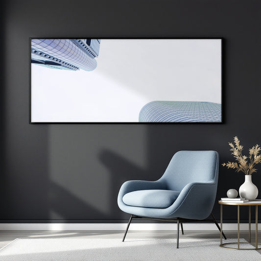 A large framed wall art print hangs on a dark charcoal wall above a light blue armchair, with a small side table and vase in the room. The artwork shows a minimalist architectural scene with blue curved building forms on a white background.