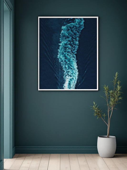 A framed wall art print showing an aerial view of a turquoise boat wake on deep blue water, displayed on a dark teal wall above a light wood floor next to a potted plant.