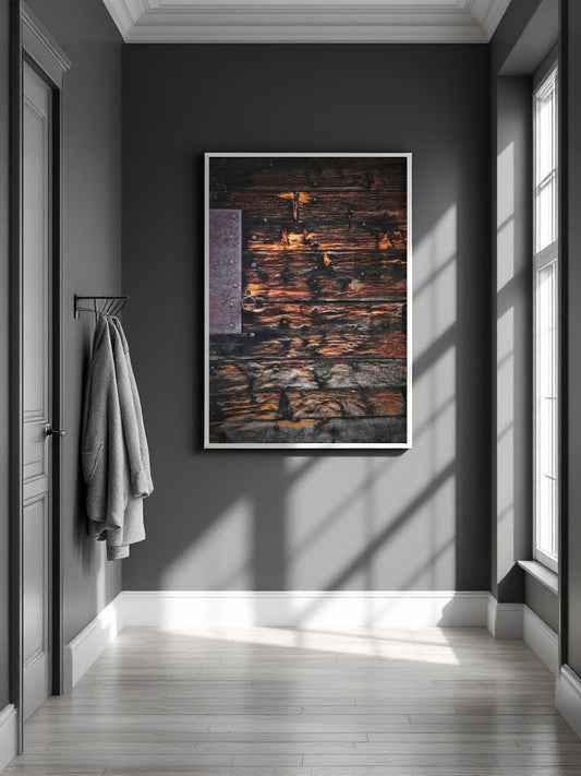 A framed abstract art print featuring textured dark wood grain with warm brown, orange, and black tones, hanging on a dark gray wall in a modern hallway with light-colored wood floors and a window casting shadows.