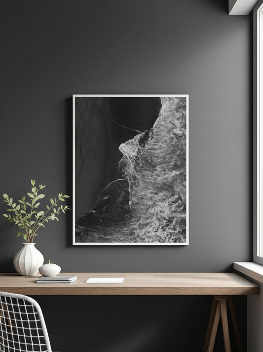 A framed black and white abstract art of ocean waves print hanging on a dark gray wall above a wooden desk with a vase and plant.