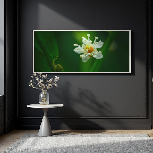 A framed wall art print showing a close-up white flower with a yellow center on a dark green background, displayed above a small round side table with a vase of dried stems against a dark charcoal wall.