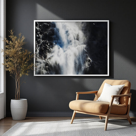 A large framed black and white waterfall art print hangs on a dark charcoal wall in a modern living room setting with a tan leather armchair and a potted plant.
