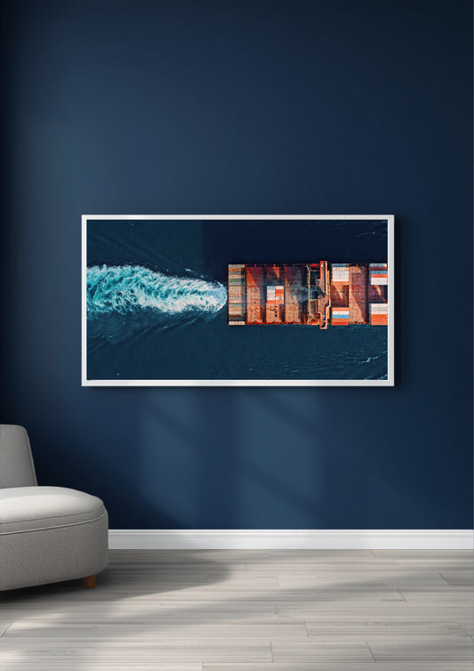 A framed horizontal art print depicting an aerial view of a cargo ship leaving a white wake on dark blue water, displayed on a dark navy blue wall above a light gray floor and a light gray chair.