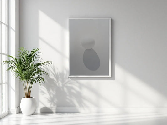 A minimalist framed wall art print with abstract gray shapes hangs on a white wall in a bright room with natural window light and a potted plant nearby.