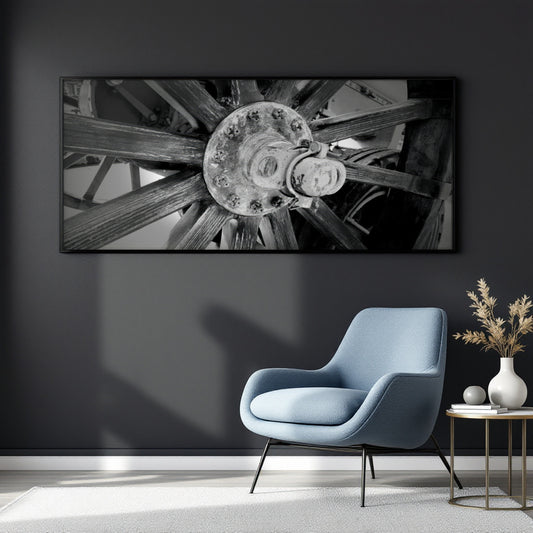 A large black and white framed wall art print depicting a close-up of a rustic wooden wheel hub, displayed on a dark gray wall above a light blue modern armchair and a small side table with a vase.