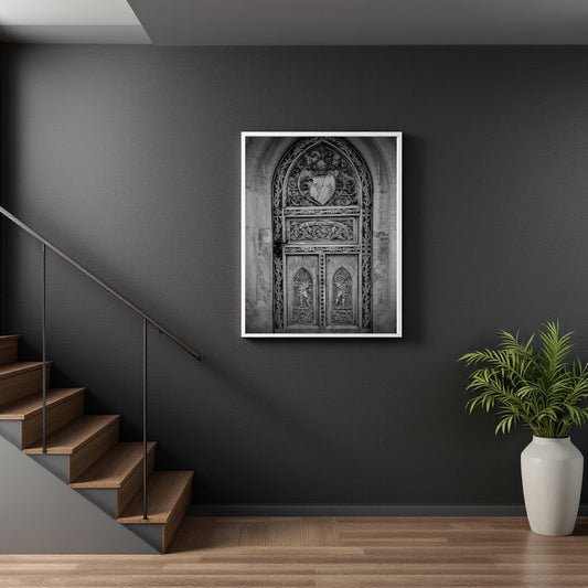A framed black and white art print of an ornate gothic-style door, displayed on a dark gray wall in a modern interior setting with a wooden staircase on the left and a potted plant on the right.