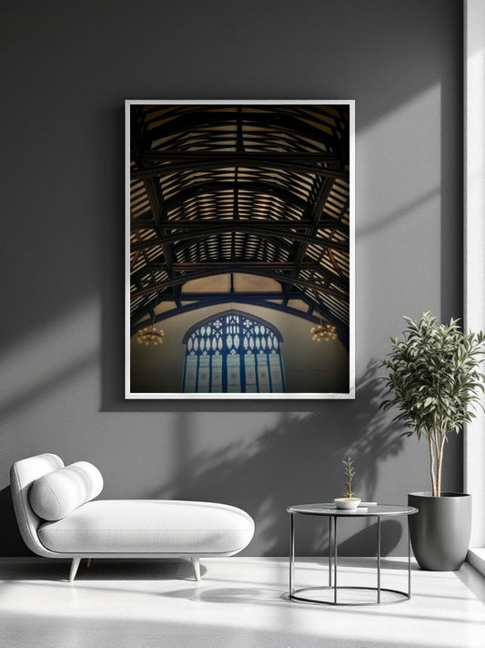 A framed vertical art print depicting an industrial stone arch ceiling and a blue stained-glass window, displayed on a dark gray wall in a modern living room setting with a white lounge chair, side table, and potted plant.