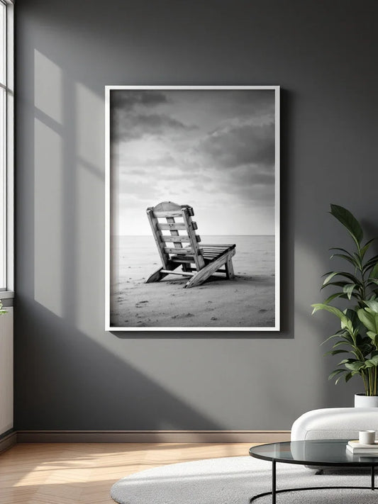 A framed black and white photograph of a wooden beach chair on a sandy shore under a cloudy sky, displayed on a dark gray wall in a modern living room setting.