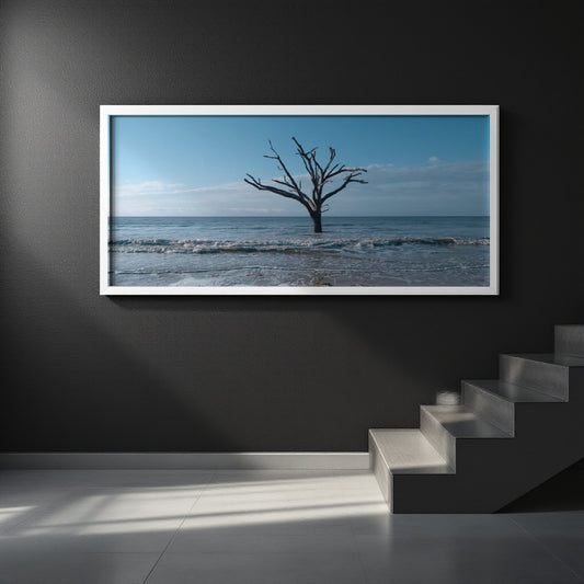 A framed horizontal art print depicting a lone bare tree standing in the ocean under a blue sky, mounted on a dark gray wall above a staircase.