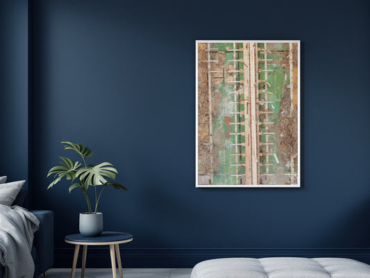 A framed abstract art print featuring textured vertical panels in green, beige, and brown tones, displayed on a dark navy blue wall in a modern bedroom setting next to a potted plant and a white tufted bench.