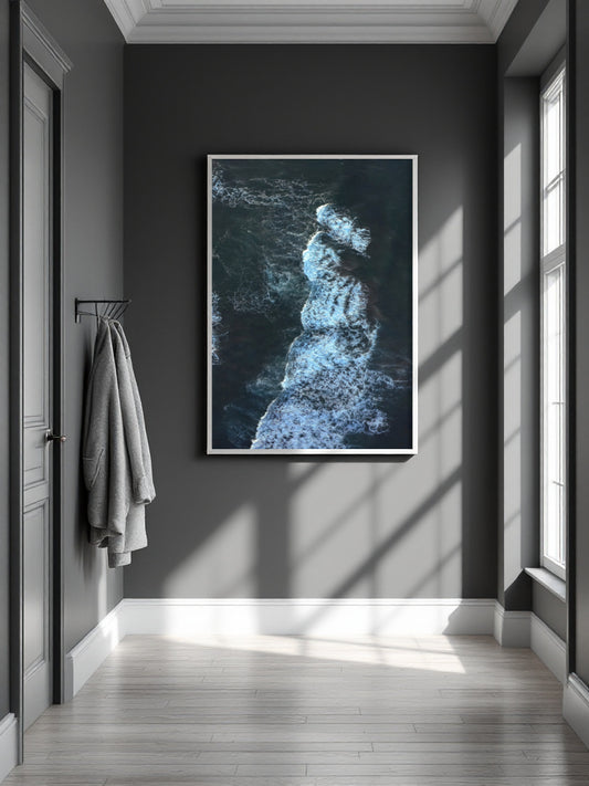 A framed vertical art print depicting a dark blue and white ocean wave crashing, displayed on a dark gray wall in a modern hallway with light wood floors and a hanging gray towel.