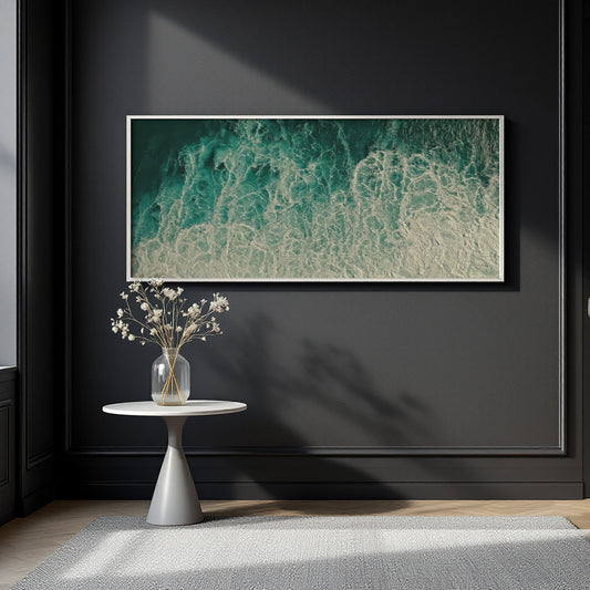 A framed abstract wall art print showing teal and white ocean waves, displayed on a dark charcoal wall above a small white side table with a vase of dried flowers.