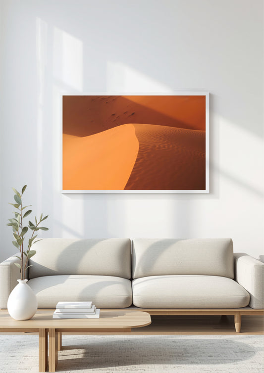 A framed wall art print showing warm orange and tan sand dunes, displayed above a light beige sofa in a bright, modern living room.