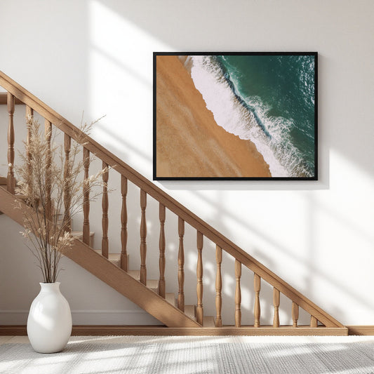 A framed square wall art print showing an aerial beach scene with turquoise ocean waves and golden sand, hung on a white wall above a wooden staircase in a bright interior.