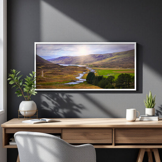 A framed landscape art print depicting a scenic valley with a winding river, green fields, and mountains under a bright sun, displayed on a dark gray wall above a wooden desk with plants and a mug.