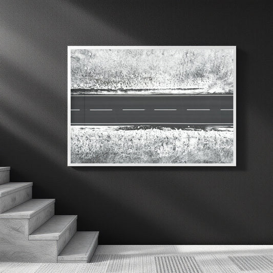 A framed black and white art print depicting an aerial view of a road cutting through textured fields, displayed on a dark wall above a staircase.