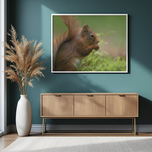 A framed wall art print showing a close-up photograph of a brown squirrel in green foliage, displayed on a dark teal wall above a light wood console table with a white vase of dried grass nearby.