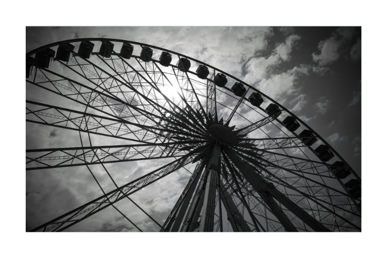 Black and white photograph of a large Ferris wheel viewed from below against a cloudy sky, presented with a white border.