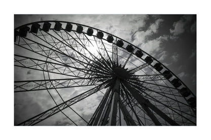 Black and white photograph of a large Ferris wheel viewed from below against a cloudy sky, presented with a white border.
