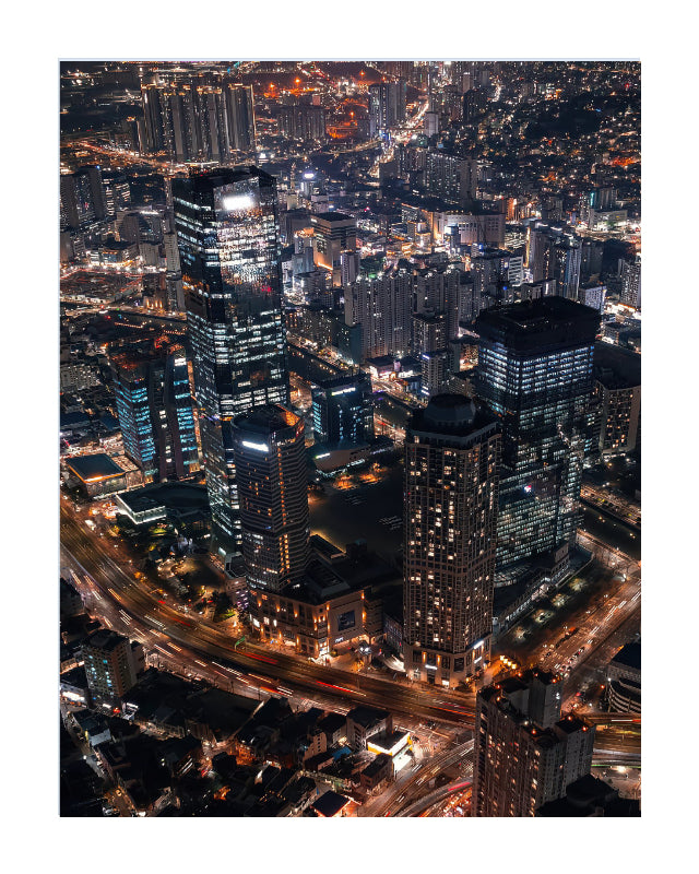 Vertical Aerial night photograph of a dense city skyline with illuminated skyscrapers, glowing windows, and light trails from roads and vehicles, presented with a white border.